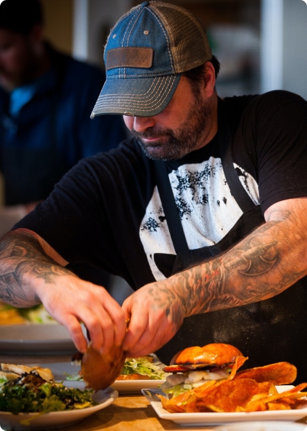 En man med en blå keps, svart t-shirt och tatueringar på armarna förbereder hamburgare med tillbehör på tre tallrikar.