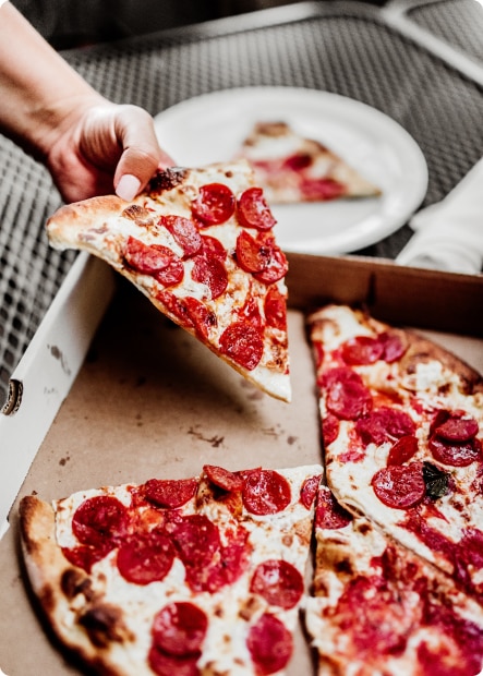 En person plockar upp en pizzaslice med salamiskivor ur en vit pizzakartong.
