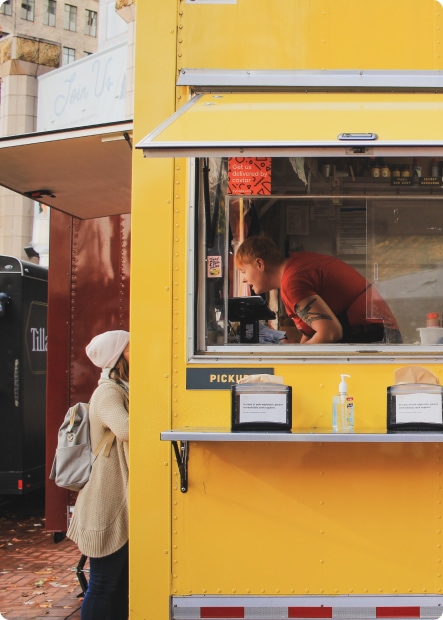 En kvinna i vitt beställer mat från en gul foodtruck och genom fönsterrutan ser man en man i röd t-shirt som tar emot beställningen.