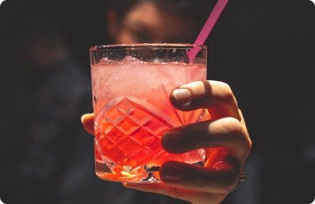 En person håller upp ett elegant glas med en röd drink med isbitar och ett rosa sugrör i.