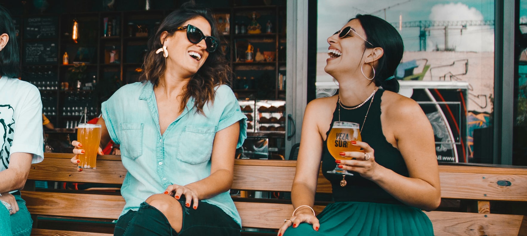 Två kvinnor sitter med varsin öl på en uteservering och skrattar.